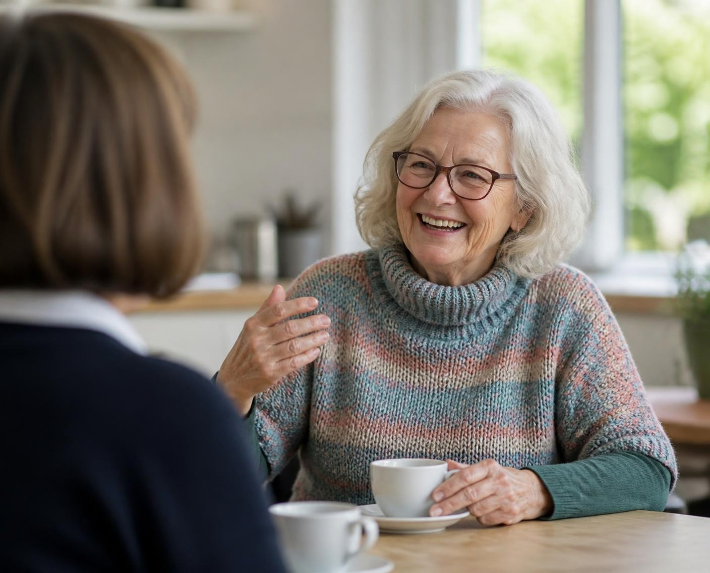 An older woman in a striped sweater laughs with someone across a table
